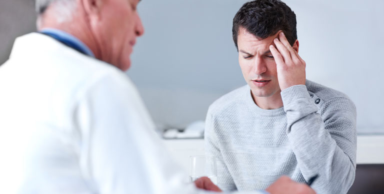 Young man rubbing his forehead while his doctor is writing out a prescription